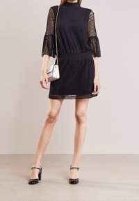 Vestido negro con un patrón texturizado, escote alto y mangas de tul acampanadas. Combinado con zapatos de tacón negros y un pequeño bolso blanco.
