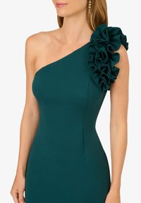 Tyrkysové šaty na jedno rameno s přiléhavým střihem. Obsahují velký volán na rameni. Hladká textura látky.