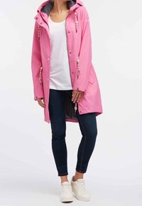 Nainen, jolla on päällään vaaleanpunainen hupputakki, valkoinen t-paita, tummat skinny-farkut ja valkoiset lenkkarit, seisoo yksinkertaista taustaa vasten.