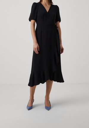 Femme portant une robe portefeuille noire avec manches bouffantes et un ourlet à volants, assortie à des talons bleus à bout pointu, debout sur un fond uni.