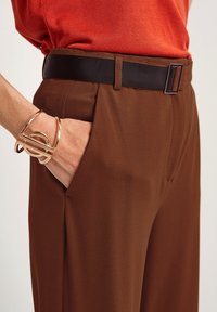Bruna byxor med en texturerad tyg, prydda med ett svart bälte och en hand dekorerad med ett guldarmband, placerad i fickområdet.