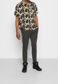 Maglietta nera a maniche corte con un motivo geometrico crema e giallo, abbinata a pantaloni grigi slim-fit e sneakers nere alte.