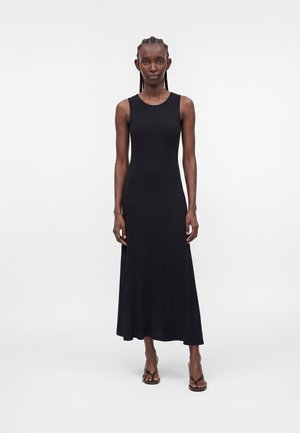 Femme debout, droite, portant une robe noire sans manches longueur cheville et des talons noirs à brides sur un fond blanc uni.