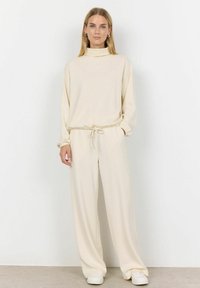 Krémový súprava loungewear pozostávajúca z topu s vysokým golierom a dlhými rukávmi, a nohavíc s nastaviteľným pásom. Jemný materiál s voľným strihom.