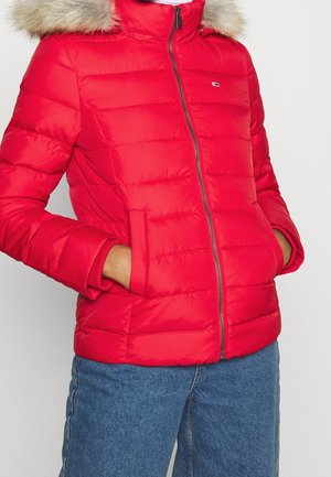 Personne portant une veste matelassée rouge avec une capuche bordée de fourrure et les mains dans les poches, associée à un jean en denim bleu.