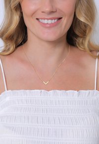 Guld kæde med en V-formet vedhæng, der har små klare accenter. Båret over en hvid tekstureret top med rynkede detaljer.
