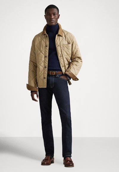 Karmelowa pikowana kurtka z brązowym kołnierzem z sztruksu, granatowy golf, ciemnoniebieskie dżinsy i brązowe mokasyny. Posiada guziki i boczne kieszenie.