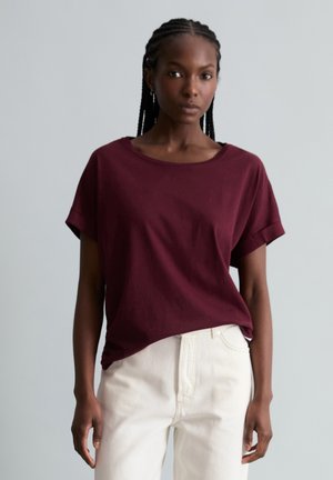 Vrouw met gevlochten haar, een losse bordeauxrode shirt met korte mouwen en hooggesneden gebroken witte broek, tegen een effen grijze achtergrond.