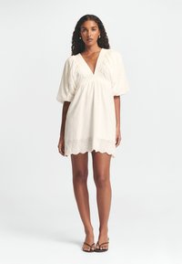 Weißes Baumwollkleid mit tiefem V-Ausschnitt, Puffärmeln und gewelltem Saum. Verziert mit floraler Stickerei und einem gesmokten Taillenbereich.