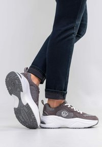 Person in dunklen Jeans und grauen sowie weißen Giesswein Sneakern, ein Fuß flach und der andere angehoben, um die Sohle zu zeigen.