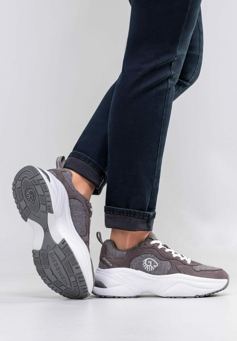 Person in dunklen Jeans und grauen sowie weißen Giesswein Sneakern, ein Fuß flach und der andere angehoben, um die Sohle zu zeigen.