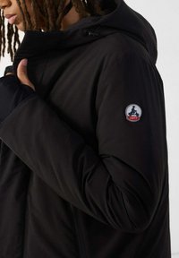 Persoon die de kraag aanpast van een zwarte hoodie met een ronde JOTT-logo patch op de bovenarm.