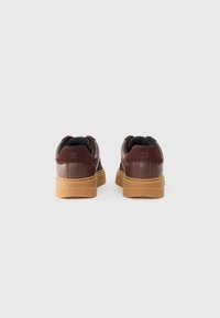 Brown leather sneakers feature a rounded shape, padded collar, black lining, and a gum outsole, with “HUGO” embossed on the heel.