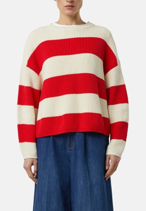 Woman wearing a red and white wide-striped knit sweater and loose blue denim pants, standing against a plain background.