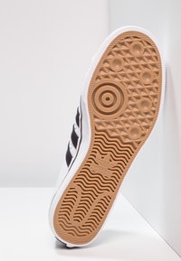 Vit sneaker med svarta ränder, gummisula med ett strukturerat gummimönster, och en logotyp nära hälen, sedd från undersidan.