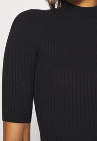 Haut en tricot noir côtelé avec un col montant et des manches courtes. Présente des rayures verticales et une silhouette ajustée. Texture douce.