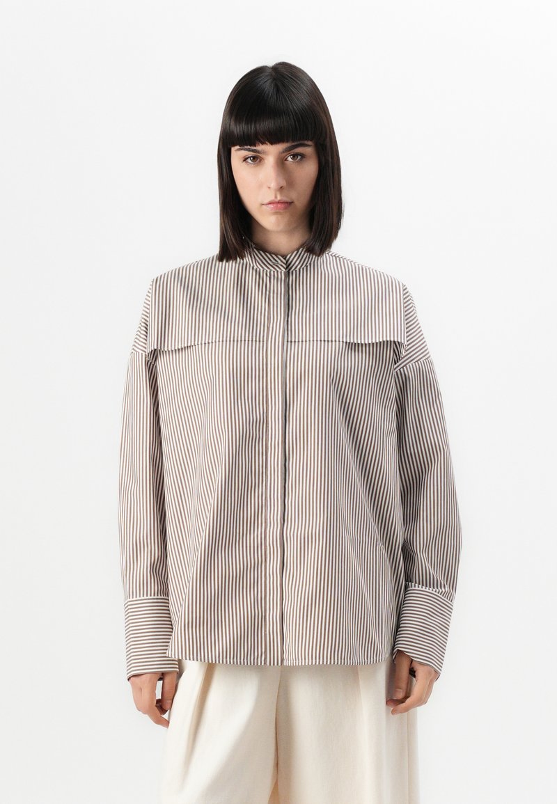 Camicia a righe nei toni beige e marrone, con colletto alto, maniche ampie e una vestibilità ampia, caratterizzata da un pannello frontale asimmetrico.