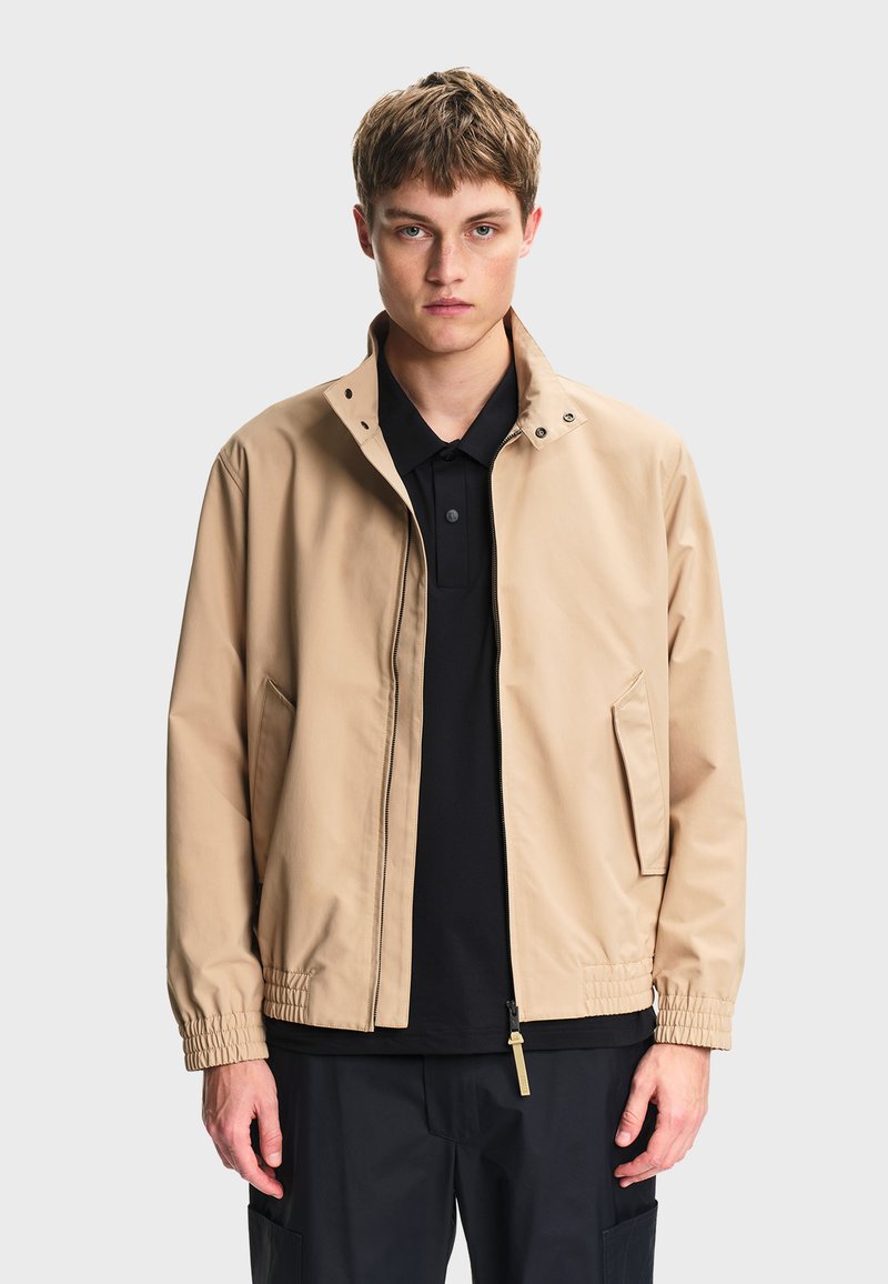 Νεαρός άνδρας που φοράει μπεζ μπουφάν με φερμουάρ και ελαστικές μανσέτες πάνω από μαύρο πουκάμισο με γιακά, στέκεται μπροστά σε έναν απλό φωτεινό φόντο.