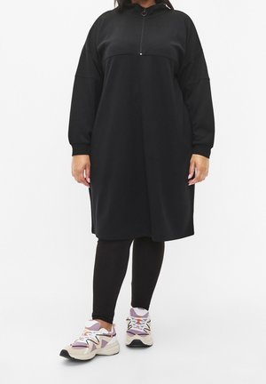 Personne portant une longue robe-pull noire zippée sur des leggings noirs et des chaussures de sport blanches, roses et violettes, debout sur un fond blanc.