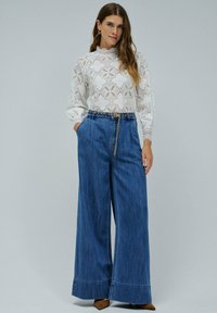 Mujer con blusa de encaje blanca de mangas largas y jeans de tiro alto de pierna ancha en azul, con un delgado cinturón de cadena dorada, de pie frente a un fondo liso.