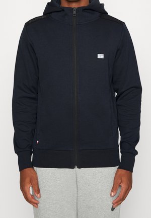 Homme portant un sweat à capuche bleu marine zippé avec un petit logo sur la poitrine et un pantalon de jogging gris, se tenant devant un fond uni.
