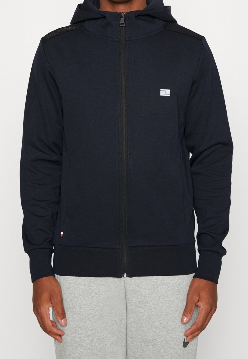 Homme portant un sweat à capuche bleu marine zippé avec un petit logo sur la poitrine et un pantalon de jogging gris, se tenant devant un fond uni.