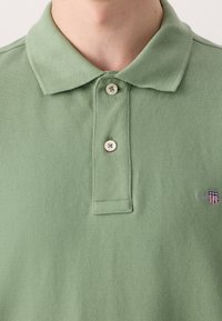 Gros plan d'une personne portant un polo vert clair avec deux boutons attachés et un petit logo brodé sur le côté droit.