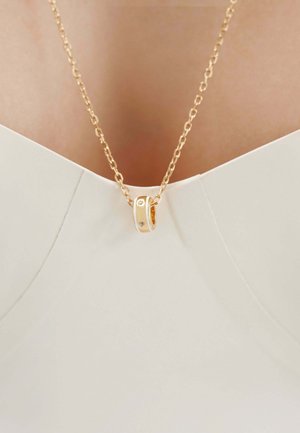 Collar de oro con un pequeño colgante circular que presenta tres diamantes incrustados, llevado con una camiseta blanca de cuello en V.