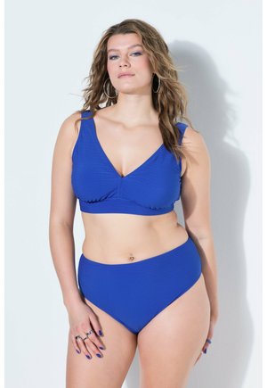 Kvinde med bølget hår iført koboltblåt ribbet bikini sæt, øreringe i hoops og ringe, stående mod en hvid baggrund.