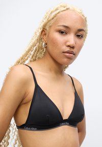 Γυναίκα με μακριά ξανθά κοτσίδια φοράει μαύρο bralette Calvin Klein με λεπτές τιράντες, στραμμένη ελαφρώς προς τα αριστερά, μπροστά σε ένα απλό φόντο.