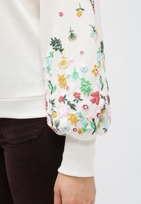 Λευκό φούτερ με πολύχρωμη κεντητή διακόσμηση από λουλούδια σε ένα μανίκι, με διάφορα φυτά και λουλούδια. Οι μανσέτες είναι ριμπ και ελαστικές.