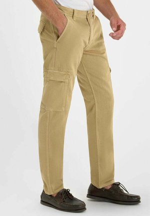 Uomo con pantaloni cargo beige e scarpe da barca marrone scuro, in piedi con la mano sinistra in tasca contro uno sfondo semplice.