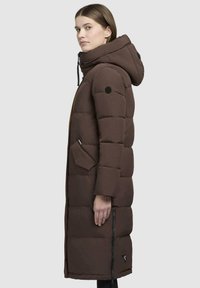 Giacca a vento marrone con cappuccio, caratterizzata da un design trapuntato, tasche laterali, accenti neri e una tasca con zip sul lato inferiore.