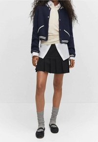 Marineblauer, verkürzter Cardigan mit weißen Streifen, über einem cremefarbenen Oberteil und einem weißen Hemd getragen, kombiniert mit einem schwarzen, plissierten Rock und grauen Socken.