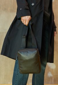 Bolso de cuero negro texturizado con forma rectangular, que cuenta con un cierre de cremallera en la parte superior y una correa ajustable. Logotipo impreso en la parte delantera.