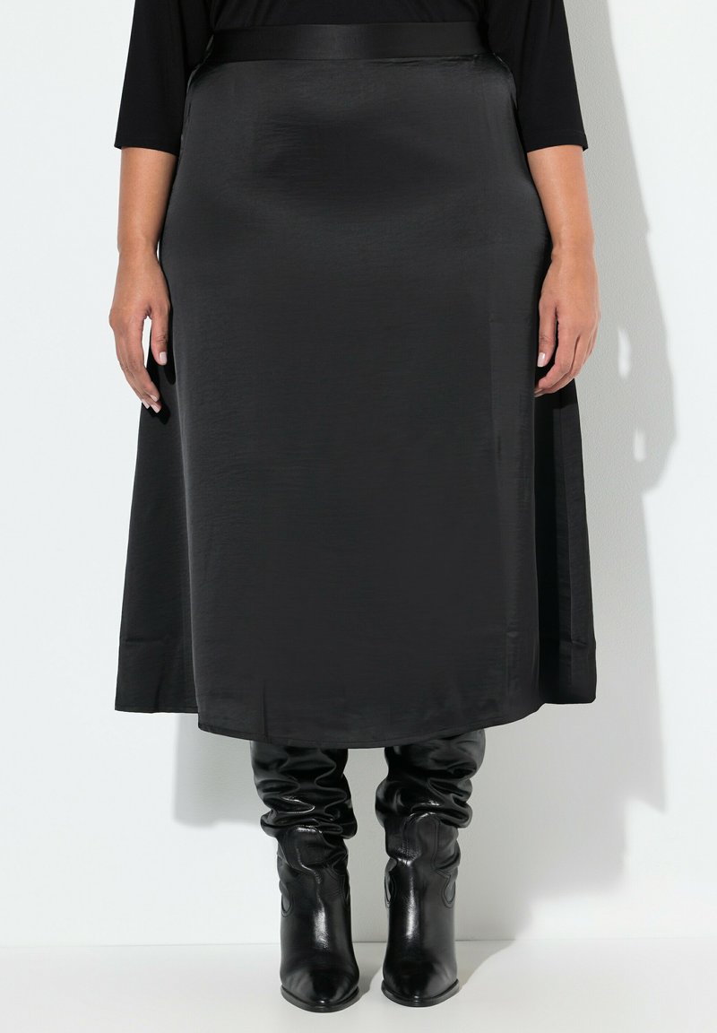 Svart satin midi kjol med slät textur, A-linjeform och sidslits, kombinerad med svarta knälånga, slappa stövlar.