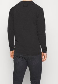 Uomo che indossa una camicia nera attillata a maniche lunghe e jeans neri, visto di spalle su uno sfondo chiaro e uniforme.
