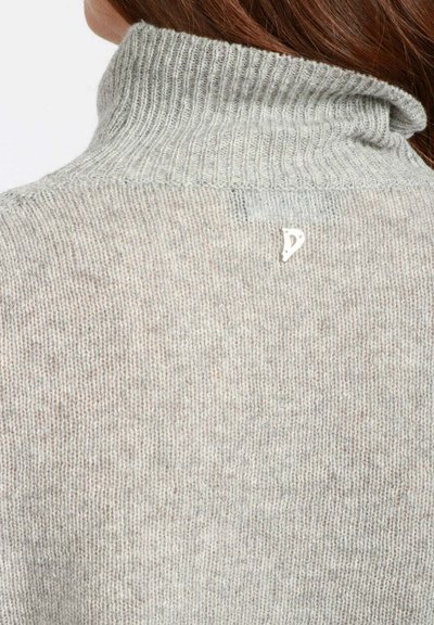 Primo piano di una persona che indossa un maglione a collo alto in maglia grigia chiara, con in evidenza il colletto posteriore e un piccolo logo bianco.