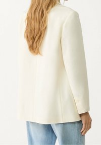 Kremowa marynarka z wyraźnie zaznaczonymi ramionami, zapinana na jeden guzik, z długimi rękawami, noszona na jasnoniebieskich dżinsach, o gładkiej fakturze materiału.