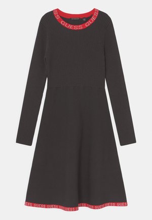 Czarna prążkowana sukienka z długim rękawem, z czerwonym wykończeniem przy dekolcie i dole, ozdobiona powtarzającym się napisem "GUESS".
