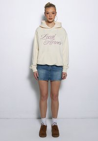 Beżowa bluza z kapturem z napisem "Local Heroes", zestawiona z dżinsową spódnicą. Modelka nosi brązowe buty i białe skarpetki, a jej włosy są związane do tyłu.