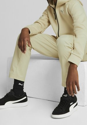 Beige urheiluhuppari, jossa on vetoketju ja logo, samaa beigen väristä housua, mustat lenkkarit, joissa on valkoisia yksityiskohtia ja logo, mustat sukat. Materiaali on teksturoitua.