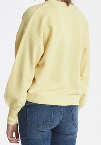 Gul tröja med avslappnad passform, sänkt axel, ribbade ärmslut och nederkant, tillverkad av mjukt material, tillsammans med blå jeans.