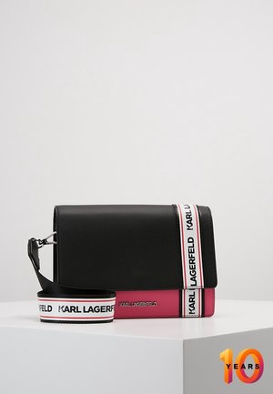 Sort og pink Karl Lagerfeld skuldertaske med hvidt mærket bånd, der ligger på en hvid overflade mod en ensfarvet baggrund.