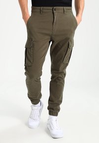 Homme portant un pantalon cargo vert olive avec des poignets élastiques et des baskets blanches, les mains dans les poches, debout sur un fond clair uni.