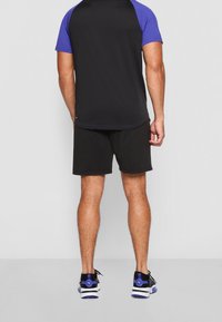 Camiseta atlética negra con mangas raglán moradas, combinada con pantalones cortos negros. Modelo que lleva zapatillas negras y azules. Diseño minimalista.
