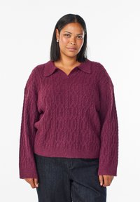 Neulepaita syvän burgundin värissä, jossa on teksturoitu kaapelikuvio, varustettuna kaarrellu kaulus ja rento malli. Yhdistetty tummaan farkkuhousuun.