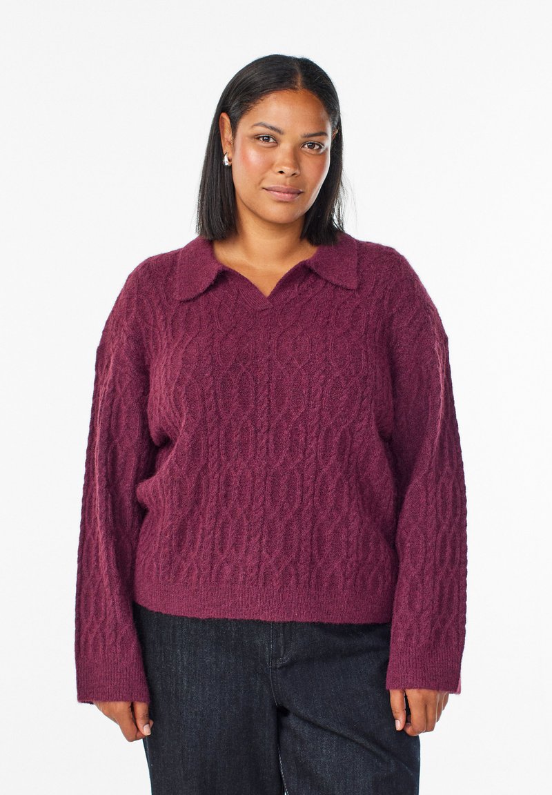 Neulepaita syvän burgundin värissä, jossa on teksturoitu kaapelikuvio, varustettuna kaarrellu kaulus ja rento malli. Yhdistetty tummaan farkkuhousuun.