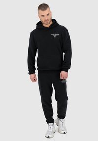 Felpa nera con cappuccio e pantaloni abbinati in tessuto morbido, con tasca frontale e dettaglio del logo. Le scarpe sono bianche con un design testurizzato.