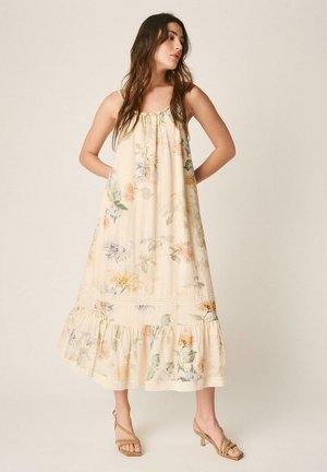 Mujer con vestido midi sin mangas color crema con estampado floral, bajo con volantes y sandalias de tacón color canela, de pie con las manos detrás de la espalda.